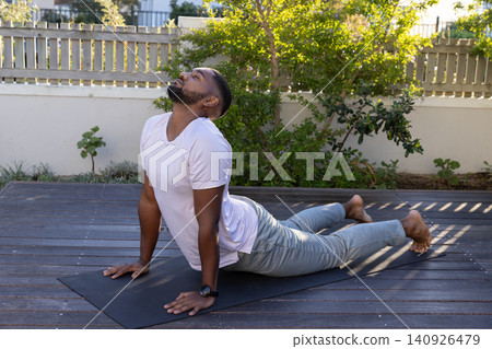 African American man doing backbend on black mat on wooden deck wearing gray sweats and smartwatch African American man doing backbend on black mat on wooden deck wearing gray sweats and smartwatch 140926479