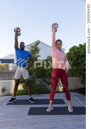 Diverse couple lifting kettlebells overhead while standing on dark mats on wooden backyard deck 140926480