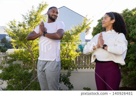 Couple standing facing each other in backyard garden holding namaste pose, wearing smartwatches Couple standing facing each other in backyard garden holding namaste pose, wearing smartwatches 140926481