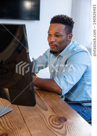 African American man sitting at wood desk working on large monitor in office, laptop cables African American man sitting at wood desk working on large monitor in office, laptop cables 140926483