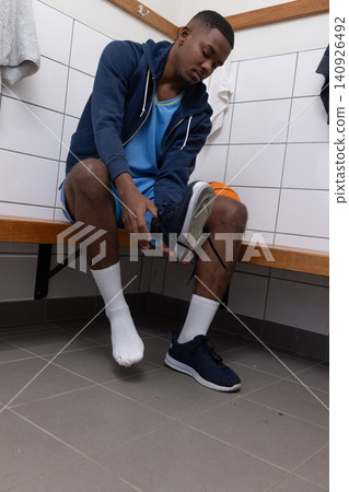 African American male sitting on wooden bench in locker room tying navy sneaker beside basketball African American male sitting on wooden bench in locker room tying navy sneaker beside basketball 140926492