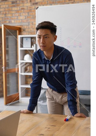 Asian man in his 30s leaning forward, pointing at whiteboard chart in office with navy shirt Asian man in his 30s leaning forward, pointing at whiteboard chart in office with navy shirt 140926494