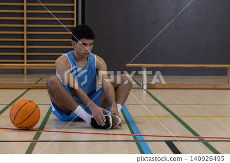 Male in blue jersey sitting on gym court reaching to hold sneaker with orange basketball nearby 140926495