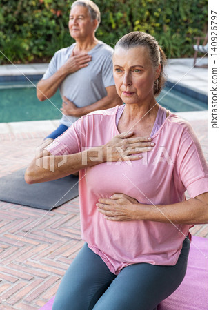Senior couple sitting on purple and dark yoga mats beside swimming pool on brick patio, breathing Senior couple sitting on purple and dark yoga mats beside swimming pool on brick patio, breathing 140926797