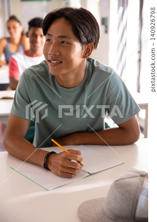 Asian teen writing with yellow pencil on spiral notebook at desk in class in light-green tee Asian teen writing with yellow pencil on spiral notebook at desk in class in light-green tee 140926798