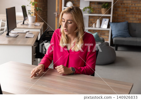 Mid30s woman wearing pink blouse sitting at wood desk in modern office pointing toward keyboard 140926833