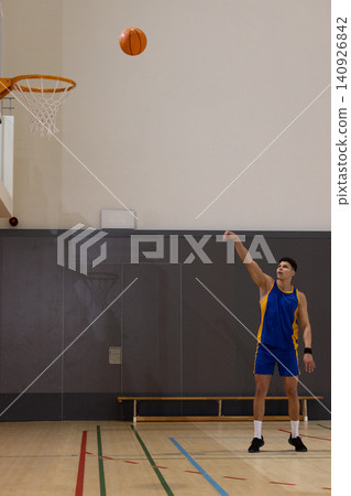 Male basketball player in blue-yellow jersey shooting ball midair in gym hoop visible, copy space Male basketball player in blue-yellow jersey shooting ball midair in gym hoop visible, copy space 140926842