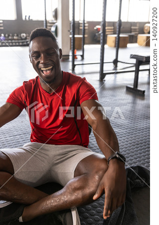 African American man wearing sportswear sitting on gym floor laughing and checking smartwatch 140927200