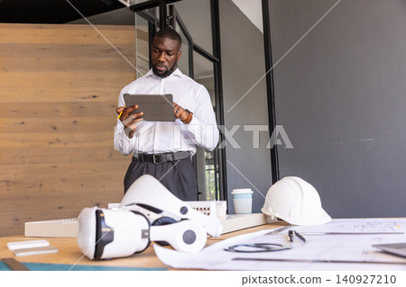 African American man standing in office reviewing plans on tablet with pencil, helmet and VR gear African American man standing in office reviewing plans on tablet with pencil, helmet and VR gear 140927210