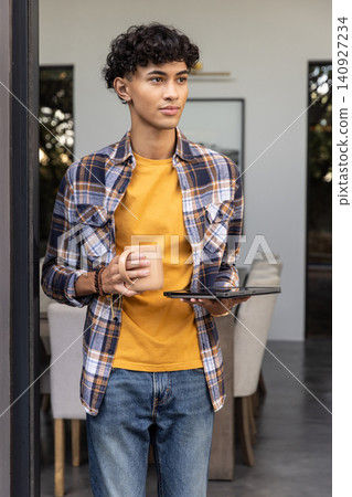Man in his twenties standing in dining area by patio holding ceramic mug and tablet Man in his twenties standing in dining area by patio holding ceramic mug and tablet 140927234