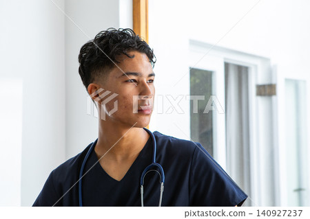 Asian male doctor standing by window frame in clinic wearing navy scrubs and stethoscope 140927237