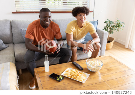 Diverse male friends sitting on sofa at home, holding football, bottle with popcorn, chips Diverse male friends sitting on sofa at home, holding football, bottle with popcorn, chips 140927243