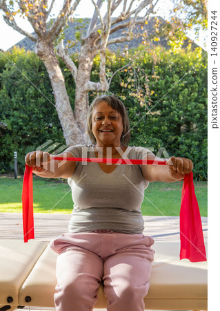 Senior african american woman pulling red resistance band, sitting on exercise bench on deck Senior african american woman pulling red resistance band, sitting on exercise bench on deck 140927244