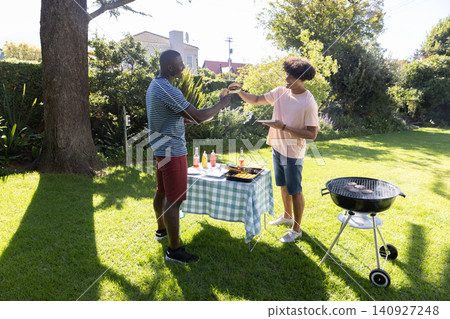 Diverse male friends clapping soda bottles, sharing burger at backyard picnic table near grill Diverse male friends clapping soda bottles, sharing burger at backyard picnic table near grill 140927248