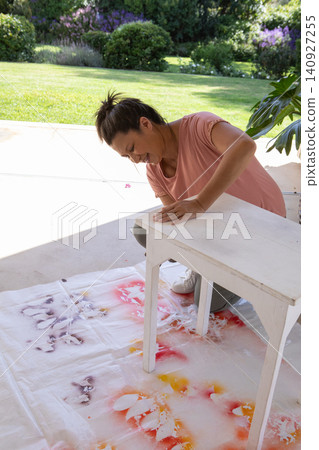 Senior woman kneeling on patio and sanding white wooden table on painted drop cloth Senior woman kneeling on patio and sanding white wooden table on painted drop cloth 140927255