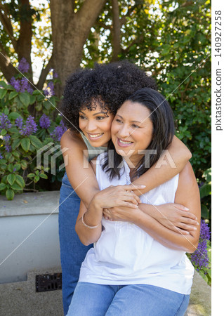 African american mother with daughter hugging on bench in yard by hedge with purple flowers African american mother with daughter hugging on bench in yard by hedge with purple flowers 140927258