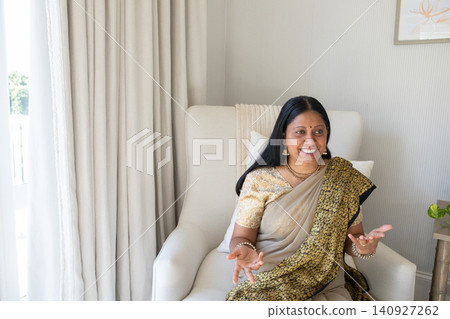 Senior indian asian woman wearing sari sitting at home on armchair with artwork Senior indian asian woman wearing sari sitting at home on armchair with artwork 140927262