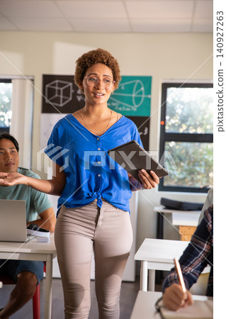 Female teacher wearing cobalt blouse and beige pants standing at front of classroom with tablet Female teacher wearing cobalt blouse and beige pants standing at front of classroom with tablet 140927263