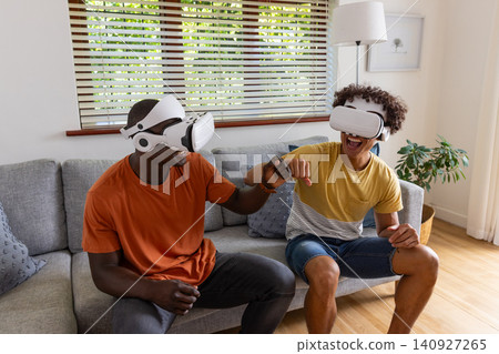 Diverse male friends bumping fists while wearing vr headsets on sofa in living room Diverse male friends bumping fists while wearing vr headsets on sofa in living room 140927265