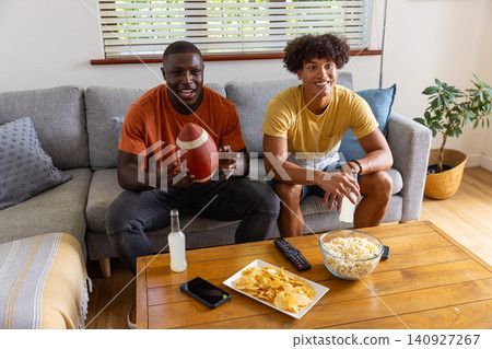 Diverse male friends holding american football, sitting on couch in living room with popcorn Diverse male friends holding american football, sitting on couch in living room with popcorn 140927267