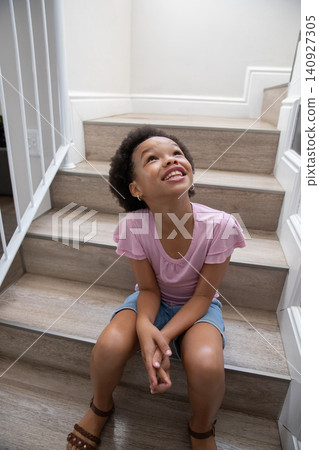 African American girl child sitting on wooden stairs inside home smiling up near metal hand railing African American girl child sitting on wooden stairs inside home smiling up near metal hand railing 140927305