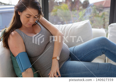 Pregnant woman sitting on sofa by window, measuring blood pressure with blue cuff Pregnant woman sitting on sofa by window, measuring blood pressure with blue cuff 140927359