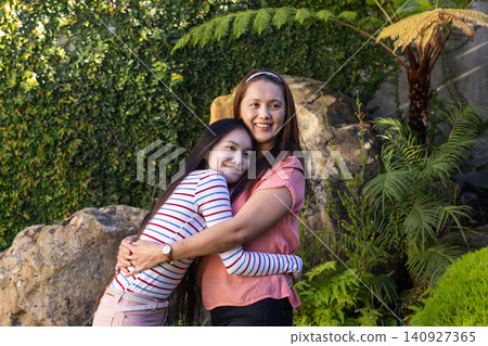 Asian mother and daughter hugging in garden with boulder, ivy wall, fern and tropical plants Asian mother and daughter hugging in garden with boulder, ivy wall, fern and tropical plants 140927365