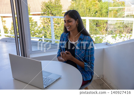 Indian woman sitting at round white table on balcony using silver laptop, smiling, copy space Indian woman sitting at round white table on balcony using silver laptop, smiling, copy space 140927416