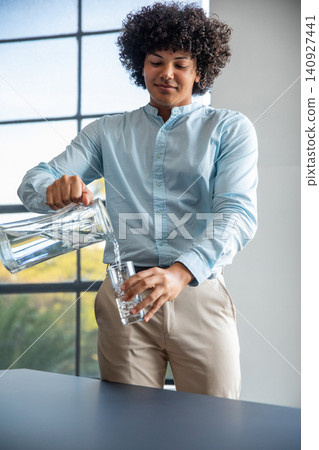 African american man in blue shirt pouring water from glass pitcher into glass in kitchen African american man in blue shirt pouring water from glass pitcher into glass in kitchen 140927441