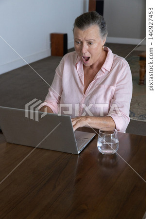 Woman typing on laptop at home on table with water glass looking surprised Woman typing on laptop at home on table with water glass looking surprised 140927453