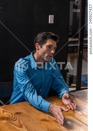 Male worker gesturing at table in meeting room, glass partition shelving Male worker gesturing at table in meeting room, glass partition shelving 140927457