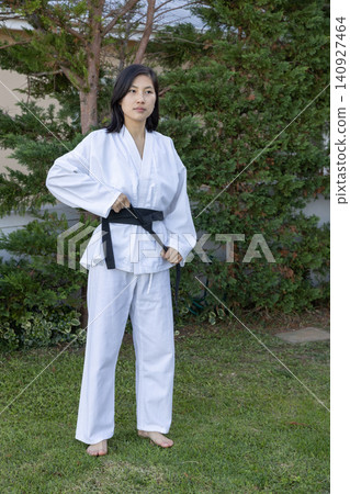 Asian woman standing barefoot on grassy lawn in backyard adjusting belt knot on white gi Asian woman standing barefoot on grassy lawn in backyard adjusting belt knot on white gi 140927464