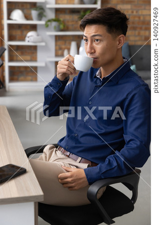 Asian man sitting at desk holding coffee mug beside smartphone in office space, copy space Asian man sitting at desk holding coffee mug beside smartphone in office space, copy space 140927469