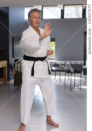 Senior man practicing martial stance barefoot in living room wearing white gi, black belt Senior man practicing martial stance barefoot in living room wearing white gi, black belt 140927472