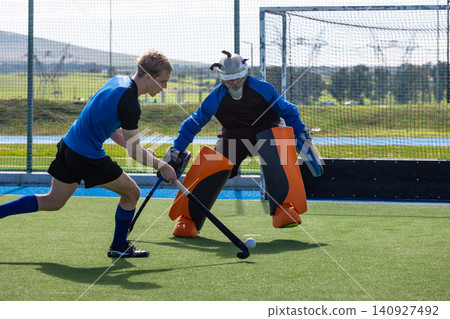 Teenage player dribbling field hockey ball as male goalkeeper in mask crouching by net on turf Teenage player dribbling field hockey ball as male goalkeeper in mask crouching by net on turf 140927492