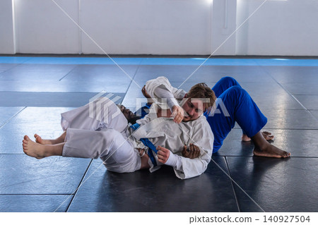 Diverse male training partners grappling, wearing gis and belts on padded mats inside dojo Diverse male training partners grappling, wearing gis and belts on padded mats inside dojo 140927504