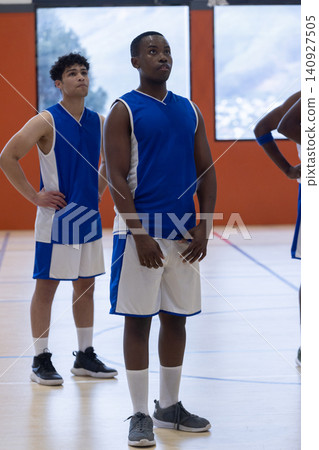Diverse teenage male teammates standing on gym court wearing basketball blue white uniforms Diverse teenage male teammates standing on gym court wearing basketball blue white uniforms 140927505
