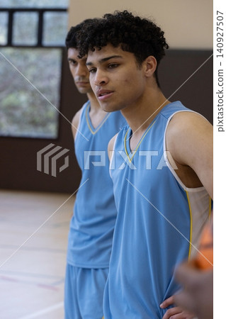Diverse male teammates in blue jerseys standing with basketball near padded wall in gym court 140927507