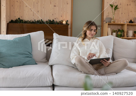 Woman wearing white hoodie sitting cross-legged on sofa near piano using tablet in candlelit room Woman wearing white hoodie sitting cross-legged on sofa near piano using tablet in candlelit room 140927520