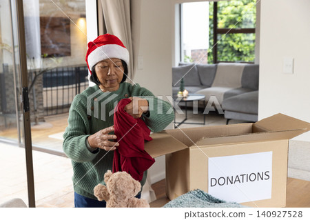 Senior Asian woman wearing Santa hat placing red garment into donation box at living room table Senior Asian woman wearing Santa hat placing red garment into donation box at living room table 140927528