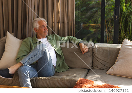 Senior man sitting on sofa at home checking smartwatch by sliding glass door, copy space 140927551
