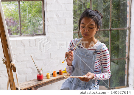 Asian teenage girl sitting on wooden bench in art studio holding paint palette, brushes, copy space Asian teenage girl sitting on wooden bench in art studio holding paint palette, brushes, copy space 140927614