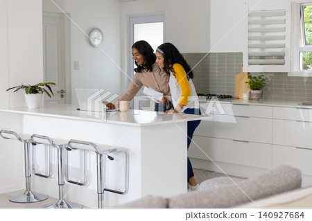 Diverse mom and teenage daughter standing at kitchen island pointing to laptop and holding papers Diverse mom and teenage daughter standing at kitchen island pointing to laptop and holding papers 140927684