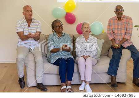 Senior friends sitting on beige couch in living room holding confetti near plant, pastel balloons Senior friends sitting on beige couch in living room holding confetti near plant, pastel balloons 140927693