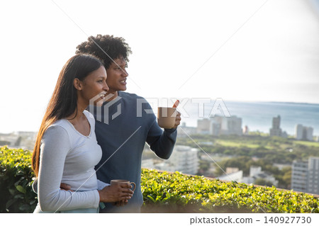Diverse couple standing on rooftop terrace by hedgerow overlooking coastal city, holding mugs Diverse couple standing on rooftop terrace by hedgerow overlooking coastal city, holding mugs 140927730