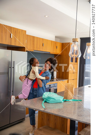 Mother lifting daughter laughing at marble countertop island in kitchen with backpack Mother lifting daughter laughing at marble countertop island in kitchen with backpack 140927737