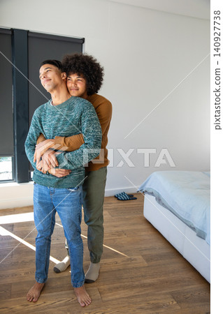 Diverse male couple embracing barefoot on hardwood in bedroom by window shade and blue bedding 140927738