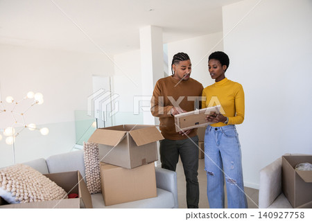 Diverse couple holding framed picture and unpacking moving boxes by light gray sofa in living room Diverse couple holding framed picture and unpacking moving boxes by light gray sofa in living room 140927758
