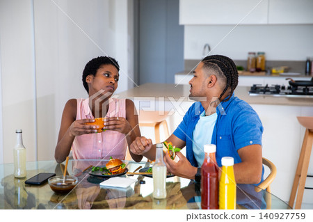 African American and Hispanic couple eating burgers at kitchen table with salad, condiment bottles African American and Hispanic couple eating burgers at kitchen table with salad, condiment bottles 140927759