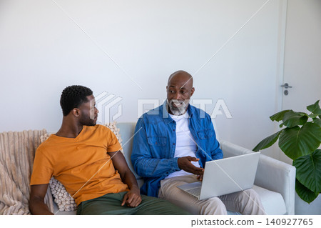African american father and son sitting on sofa in minimalist living room discussing laptop 140927765